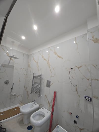 Bagno con rivestimento effetto marmo, faretti incassati nel soffitto, sanitari e un cavo in attesa che esce dalla parete.