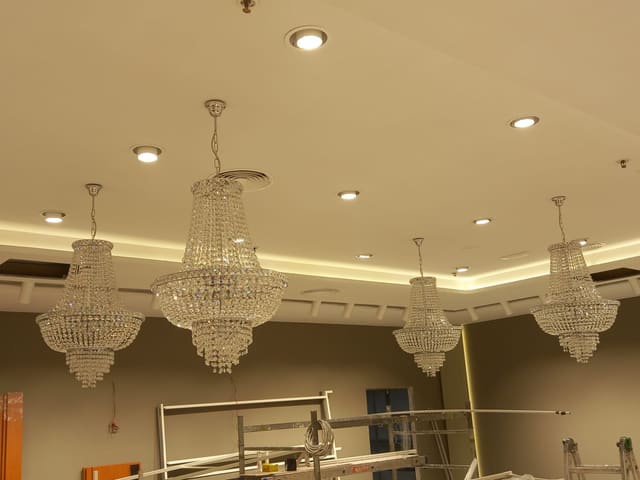 Lampadari in cristallo accesi appesi al soffitto, faretti incassati e una striscia di luce calda lungo il bordo del controsoffitto; sotto, strutture da cantiere.