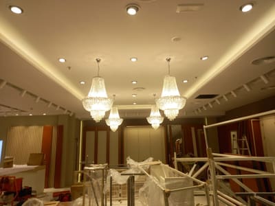 Sala con lampadari in cristallo, faretti incassati, gola luminosa e binari lungo il bordo del soffitto; in basso arredi imballati e strutture metalliche.