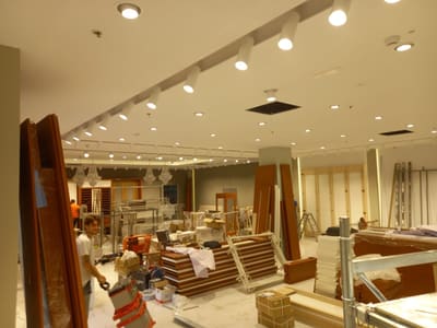 Spazio in allestimento con una fila di proiettori bianchi su binario e faretti incassati; persone al lavoro tra pannelli in legno e materiali imballati.